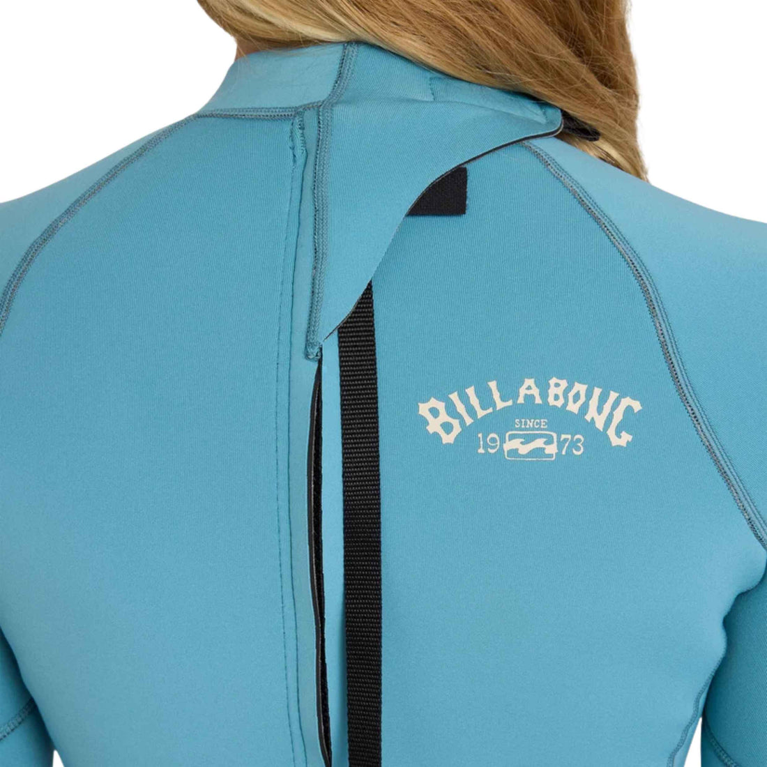 Close-up back view of a person wearing a light blue Billabong wetsuit, showing the zipper, shoulder seam, and white Billabong logo printed on the upper back.