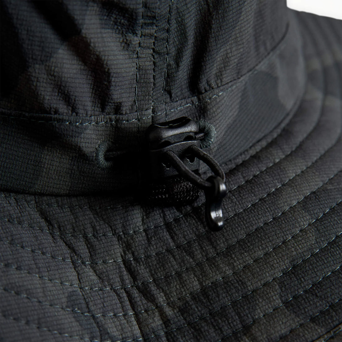 Close-up of a black camouflage bucket hat showing the textured fabric, stitching, and a black plastic drawstring toggle on the brim.