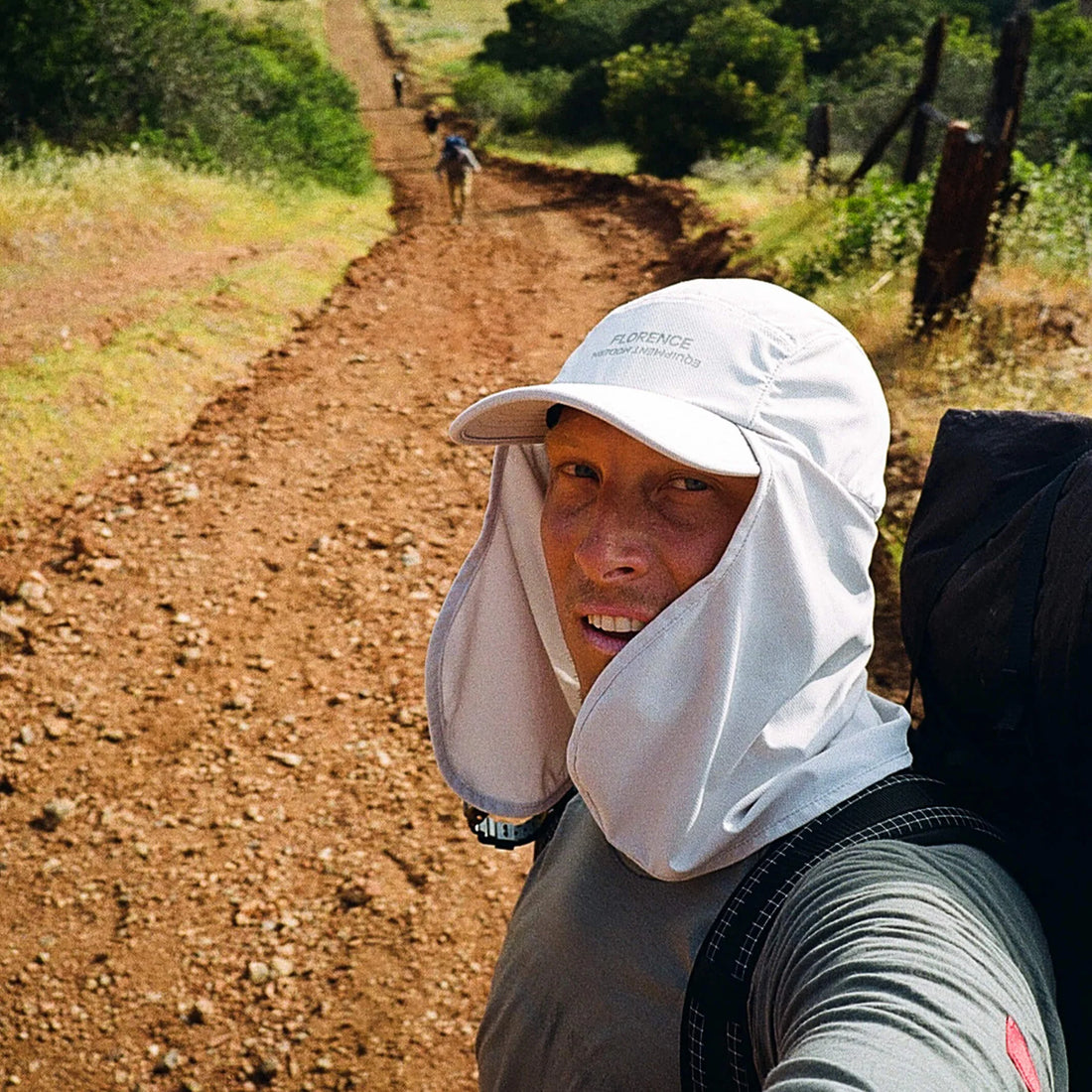 A close-up selfie of a person hiking on a dirt trail, wearing a white sun hat with a neck flap and a gray long-sleeve shirt, with a backpack visible on the right side.