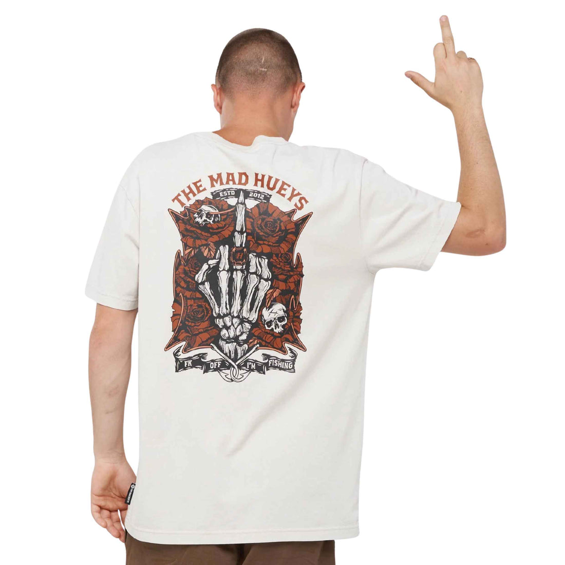 A man shown from the back wearing a cream-colored t-shirt with a large graphic print on the back. The graphic features a skeleton hand holding a cross, surrounded by red roses and skulls, with the text 'THE MAD HUEY'S' arched above.