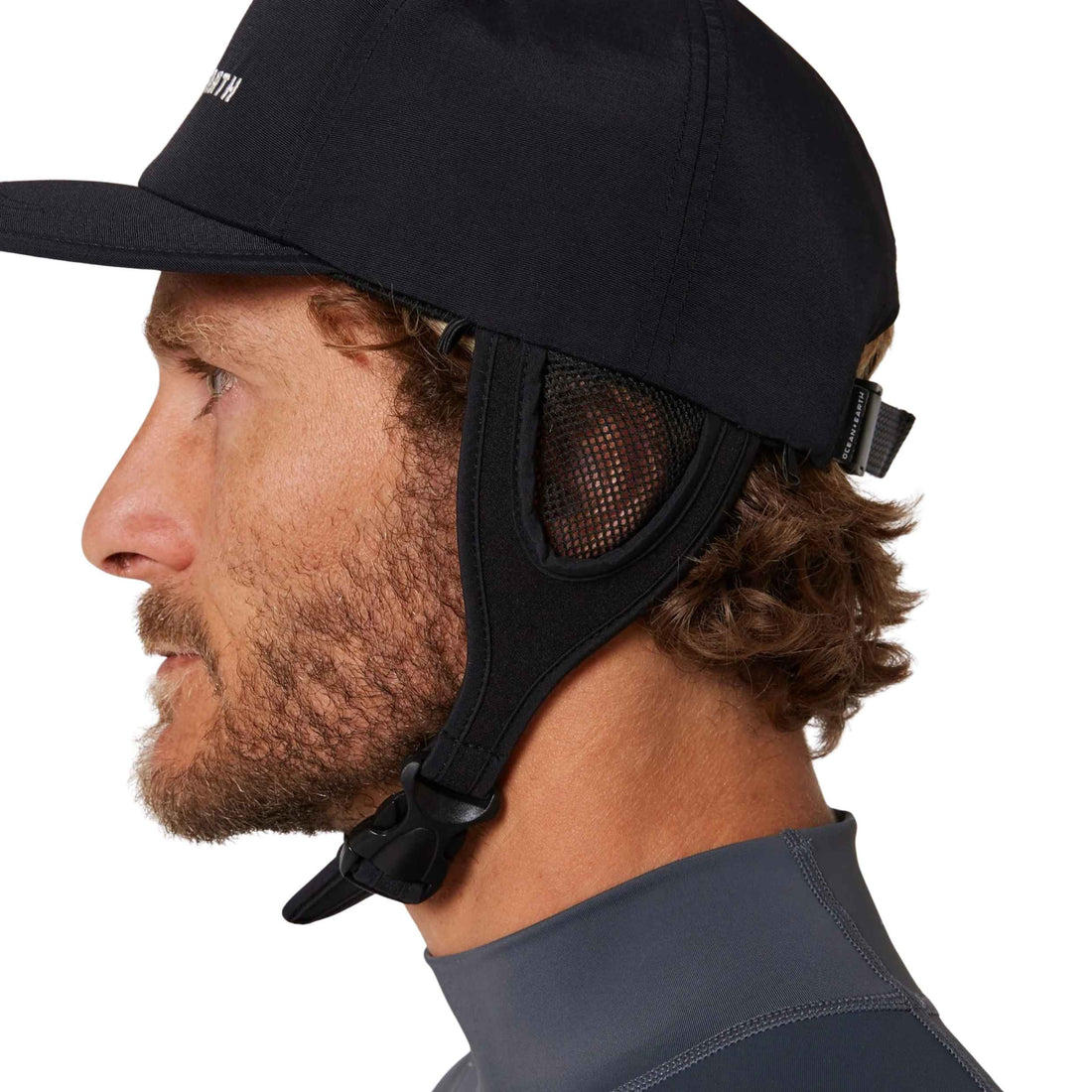 Close-up side profile of a man wearing a black wetsuit cap with an integrated mesh ear cover and adjustable chin strap, paired with a dark gray wetsuit top against a white background.
