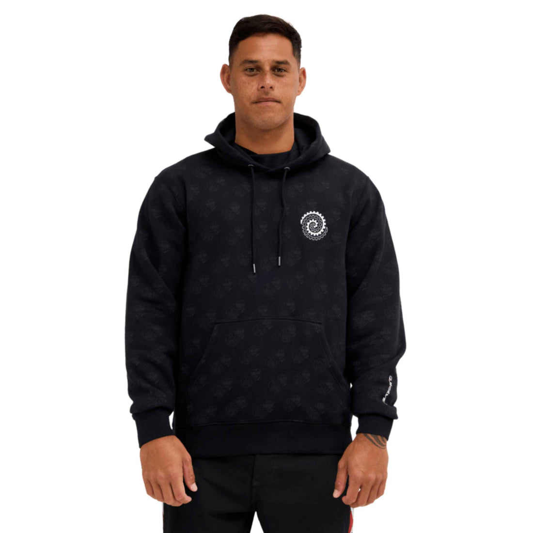 A man wearing a black patterned hoodie with a white circular logo on the left chest, photographed against a plain white background.
