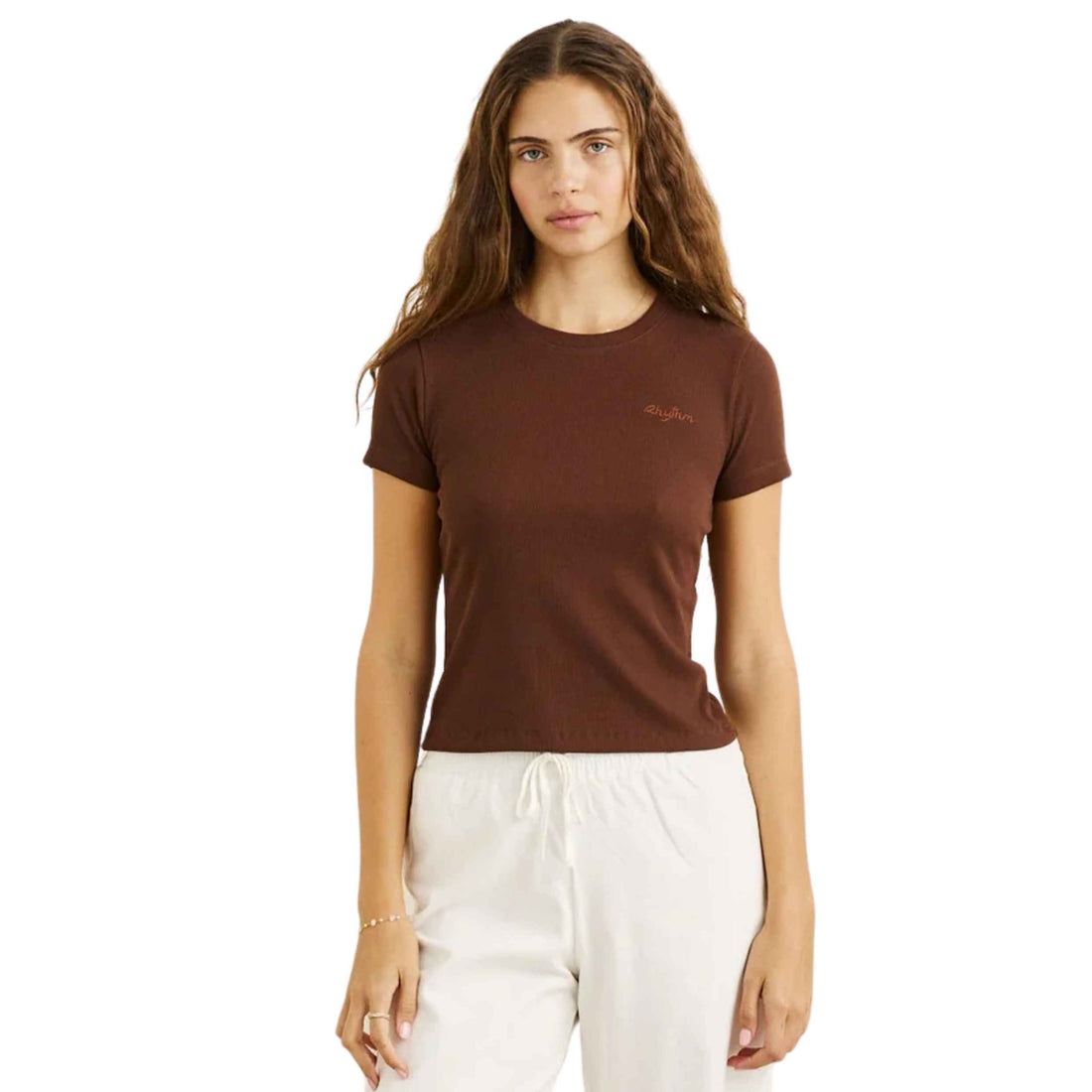 A woman modeling a brown short-sleeve crewneck t-shirt with a small embroidered logo on the left chest, paired with white drawstring sweatpants, against a plain white background.