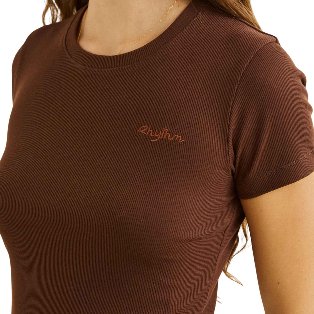 A close-up of a person wearing a brown ribbed short-sleeve crewneck t-shirt with the word 'Rhythm' embroidered in orange on the left chest, set against a plain white background.