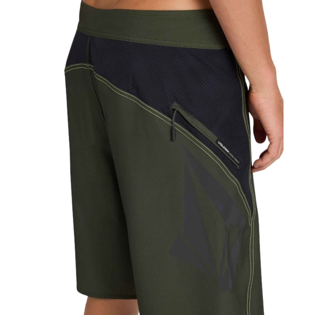 Close-up of a person wearing dark olive green and navy board shorts with a side zip pocket and drawstring, shown against a plain white background.