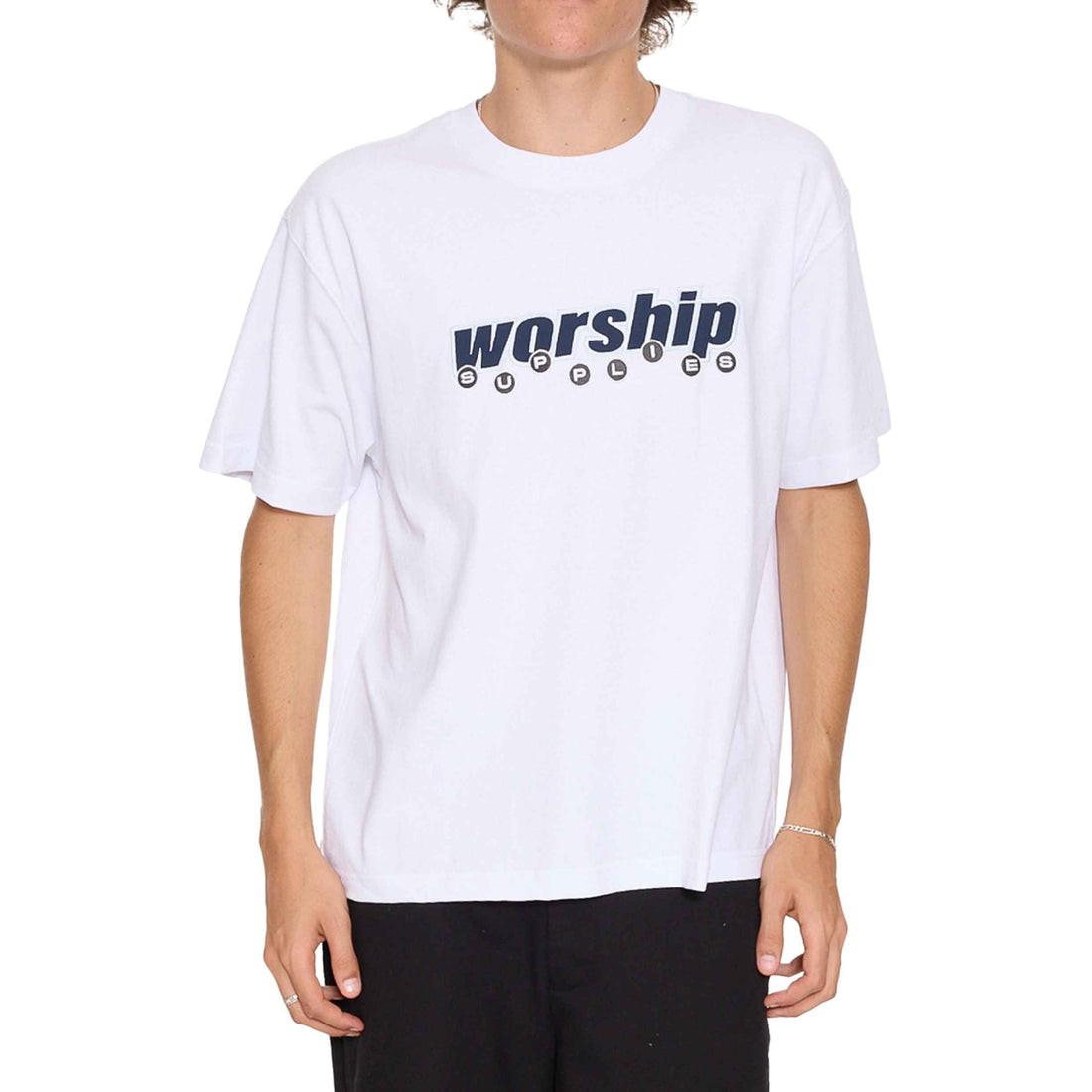 A person wearing a white short-sleeve T-shirt with a dark navy 'worship' graphic across the chest, paired with black pants, against a plain white background. Worship Line Stepper Tee White men's organic cotton t-shirt at Freeride Surf & Skate Auckland NZ