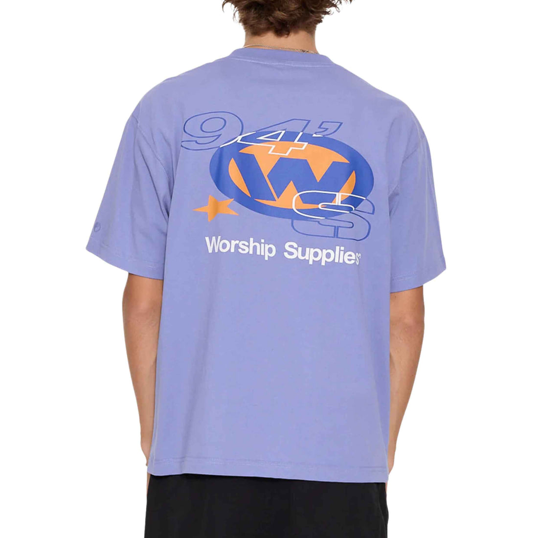 A person shown from the back wearing a light periwinkle blue t-shirt with a large graphic on the back. The graphic features a blue and orange circular 'W' logo with '94' and 'Worship Suppliers' printed in white. Worship Topper Tee Purple Haze men's organic cotton t-shirt at Freeride Surf & Skate Auckland NZ